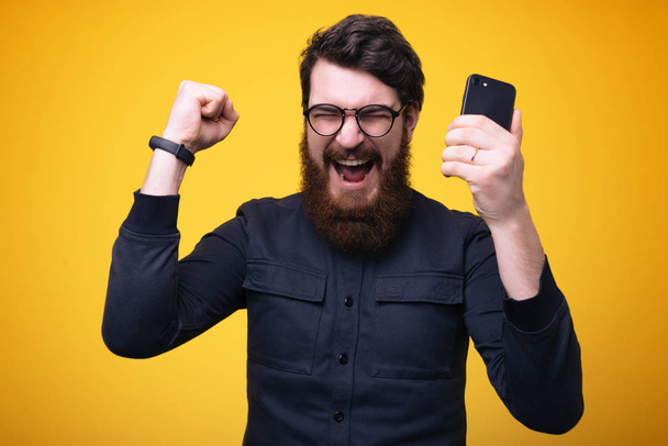 Portrait of a happy young bearded man in shirt holding mobile phone, celebrating victory, closed  eyes - Foto, immagini