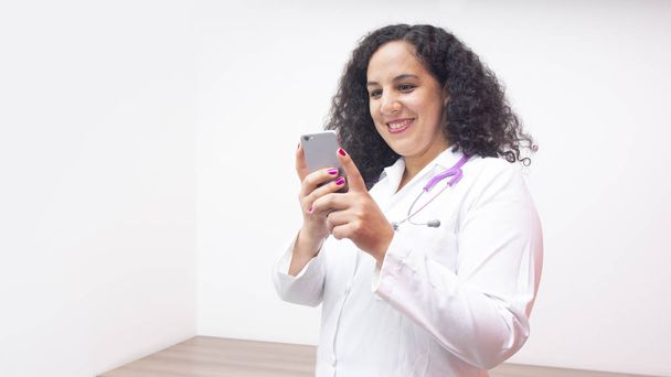女性ラテン女医笑みを浮かべて立っている白い背景の上の彼女のスマート フォンで入力する彼女の首に聴診器で彼女のオフィスで彼女の携帯電話を見て - 写真・画像