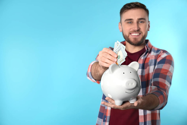 Happy young man putting money into piggy bank on color background. Space for text - Valokuva, kuva