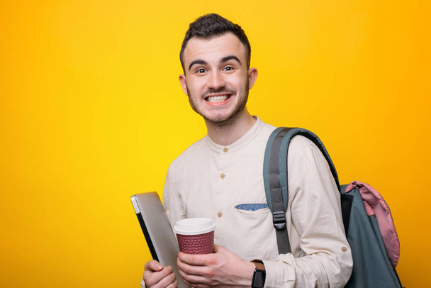 Красивий усміхнений молодий студент тримає ноутбук і чашку напою, дивлячись на камеру на жовтому тлі
 - Фото, зображення