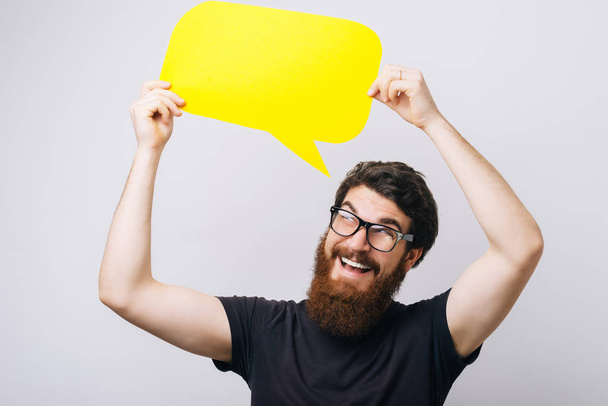 Portrait of a smiling young bearded man dressed in black shirt holding empty speech bubble over whit - Foto, afbeelding