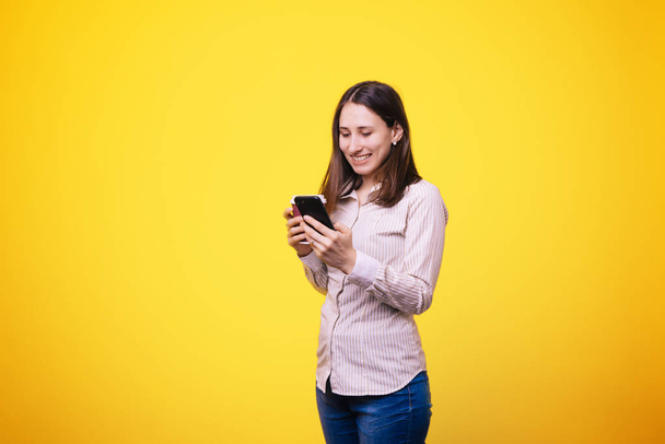 Mooie jonge vrouw met behulp van haar smartphone en drinken koffie op gele achtergrond  - Foto, afbeelding