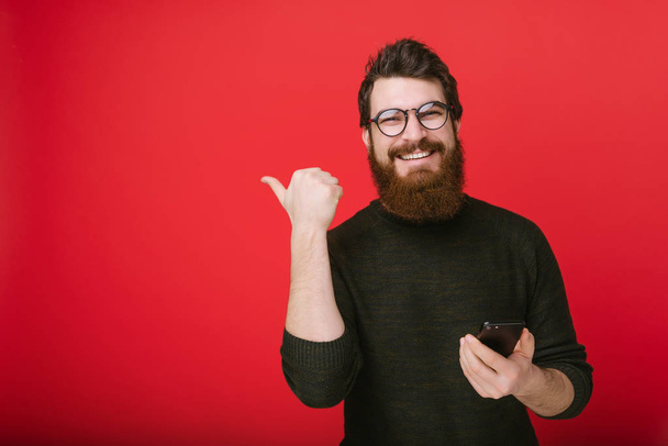 Young bearded man in glasses holding a smartphone and showing thumb up or pointing away - Photo, Image