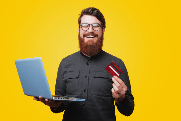 Happysmiling bearded man looking at camera while holding a laptop and showing his credit card  - Foto, Bild