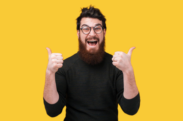 Verbaasd hipster man met baard en glazen gebaren met tumbs up - Foto, afbeelding