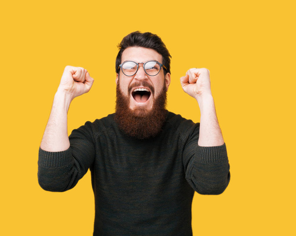 Excited bearded guy celebrate,  and gesturing with hands up standing over yellow background - Fotografie, Obrázek