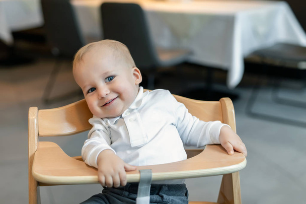 Cute little baby boy in white polo t-shirt sitting in wooden baby chair and laughing at cafe indoors. Portrait of adorable blond kid having fun at restaurant - Photo, Image