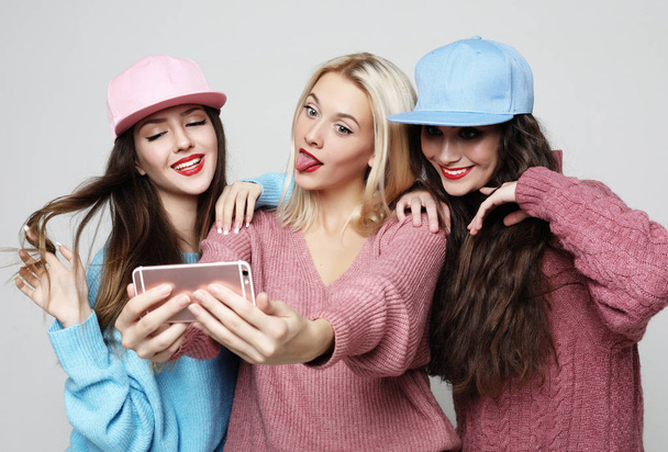 Three happy best girlfriends wearing sweaters and caps making selfie on smartphone. - 写真・画像