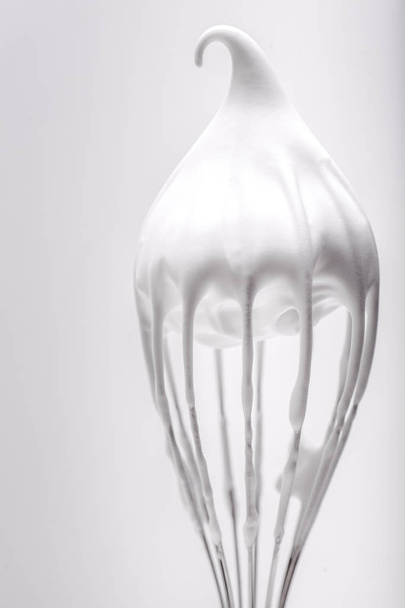 Batidor de metal con claras de huevo batidas sobre fondo blanco
 - Foto, Imagen