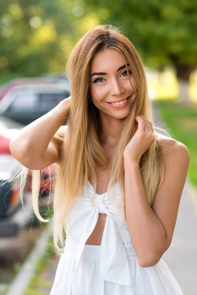 Beautiful girl with long hair straightens hair, posing in a white dress woman, tanned skin, happy smiling and joyful. Emotions of joy of happiness and outdoor recreation in city summer park. - Фото, изображение