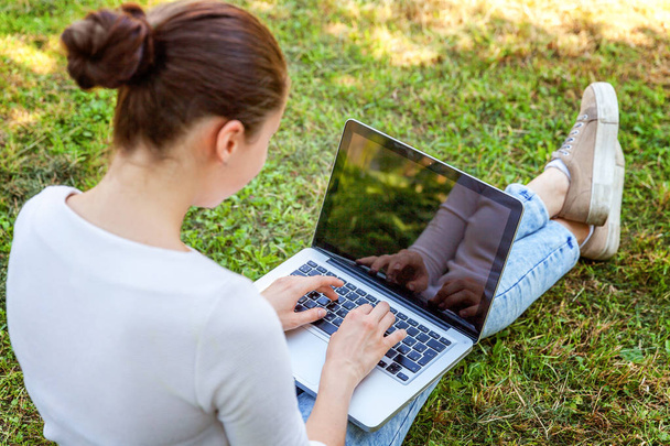 Young woman sitting on green grass lawn in city park working on laptop pc computer. Freelance business concept - Foto, Imagen