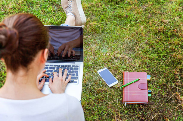Young woman sitting on green grass lawn in city park working on laptop pc computer. Freelance business concept - Zdjęcie, obraz