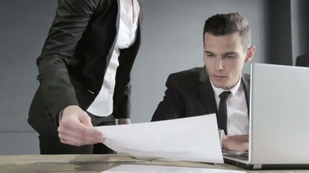 Young businessman in black suit using laptop in office, his collegue giving financial data diagram - Metraje, vídeo