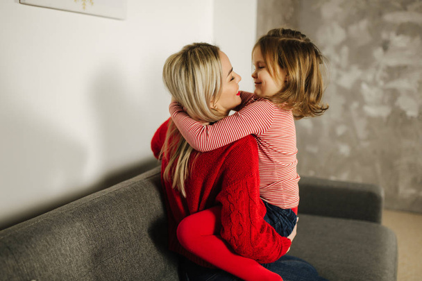 Tyttö punaisissa sukkahousuissa, äiti punaisessa puserossa. Onnellinen perhe kotona. Tytär halata hänen äitinsä
 - Valokuva, kuva