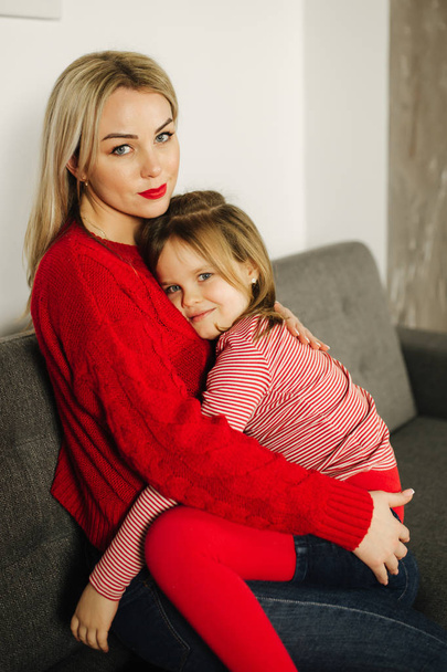 Niña en medias rojas con mamá en suéter rojo. Familia feliz en casa. Hija abrazar a su madre
 - Foto, Imagen