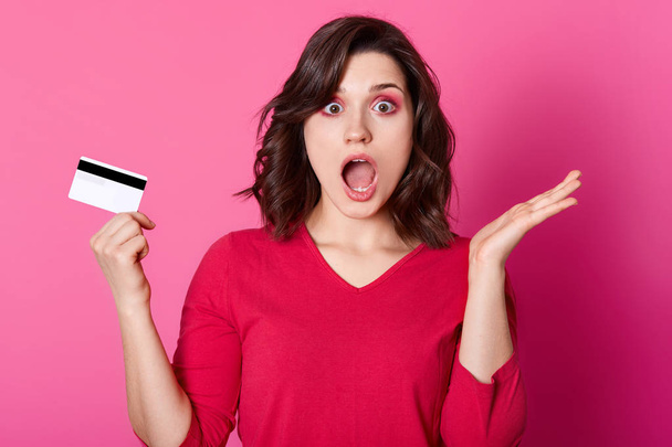 Astonished brunette woman being short of money, has no opportunity to make purchase online. Young female wears sewater stands with opened mouth isolated on pink wall. Interent and shopping concept. - Photo, Image