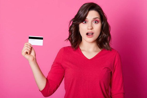 Image of beautiful shocked young woman with opened mouth isolated over rose background. Cute girl holds credit card, finds out she has no money, can not pay for purchases. Online shopping concept. - Foto, afbeelding