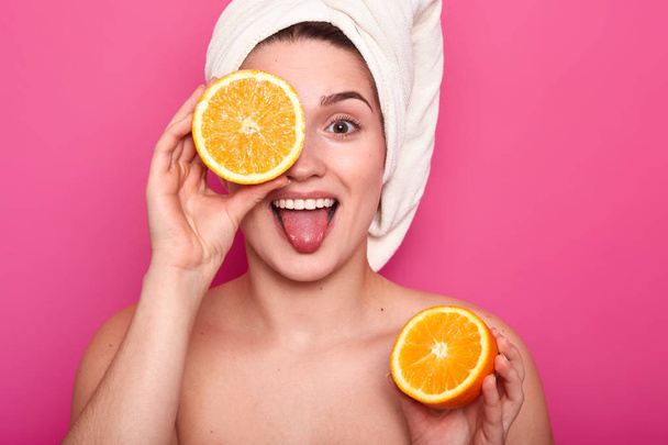 Beautiful young woman holds half of orange in her hand and covers eye with another part of fruit. Attractive lady with towel on head stands over pink background. Everyday procedure and beauty concept. - Valokuva, kuva
