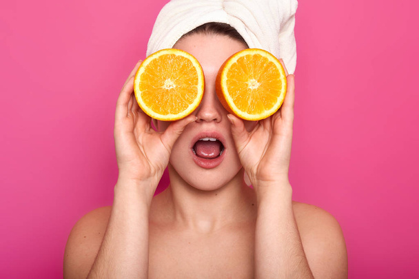 Studio shot of pleasant looking young shocked European woman covers eye with oranges, has white towel on head. Model with clear skin poses in studio isolated over pink background. Beauty concept. - Foto, immagini
