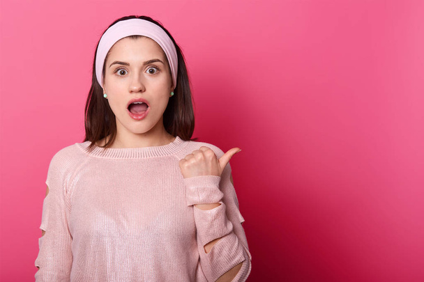 Image of european woman with dark straight hair and pink hair band, points thumb finger aside at copy space. Beautiful female stands with opened mouth, wears rose sweater against studio wall. - Photo, Image
