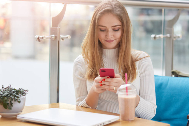 Elegant young blonde woman in white sweater sits on blue sofa in cafe, holds pink mobile phone and surfes internet or types mesage to her friend. Attractive woman drinks cocktail and check email. - Фото, зображення