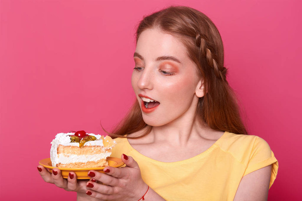 Attractive young woman with opened mouth holds plate with piece of delicious cake in hands. Brown haired lady with red manicure poses in studio isolated over pink background. Sweet food concept. - Фото, изображение
