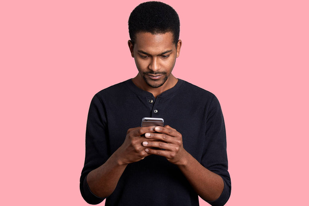 Young guy looks at the screen of smartphone with serious fecial expression. Black handsome male has important unpleasent correspondence. Boring pastime. Modern technology and Internet concept. - Valokuva, kuva