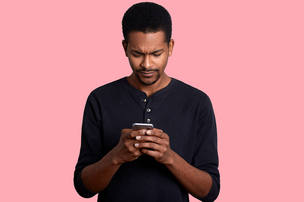 Young african man with mobile phone over rose background. Handsome dark skinned male types massege with bad news for his boss. Attractive black guy dresses casual outfit poses in photo sstudio. - Fotoğraf, Görsel