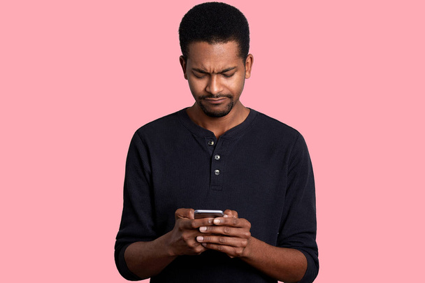 Portrait of attractive African American man with telephone in hands, surfes Internet. Handsome black male looks weather forecast for weekend with sad fecial expression. Modern technology concept. - Foto, Imagem