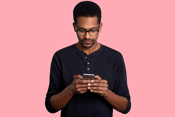 Confident young African man looks at smart phone and sends text messege, connected to wireless internet, stands against pink background. Dark skinned guy wears black shirt and glasses poses in studio. - Zdjęcie, obraz