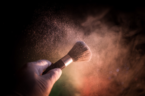 Makeup brush in hand with cosmetic powder on dark background with light and smoke. Powder splash on dark. Selective focus - Photo, Image