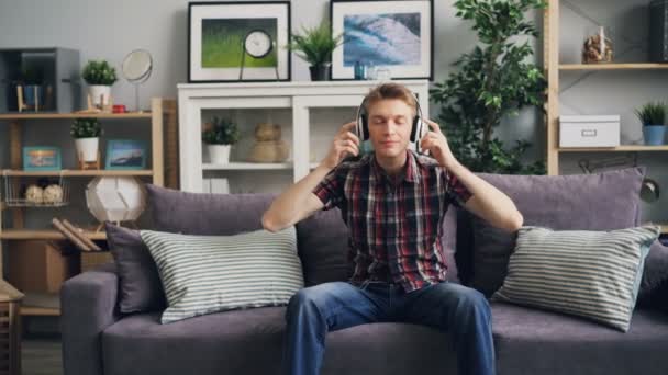 Good-looking young man in casual clothing is putting on headphones and enjoying favourite music sitting on sofa at home. Gadgets and pleasure concept. - Video