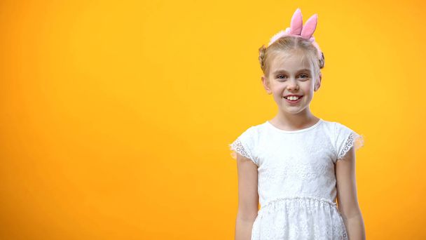 Cheerful girl in bunny ears headband smiling, isolated on orange background - Photo, image