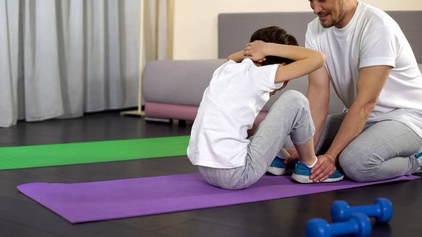 Boy doing sit-ups at home with help of father, family training, sport childhood - Foto, imagen