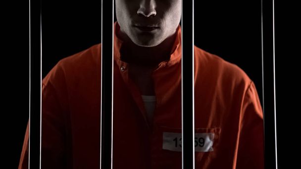Criminal in orange uniform behind prison bars, serving life sentence for murder - 写真・画像