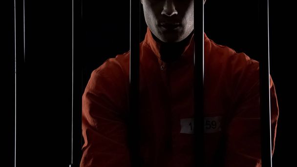 Prisoner in orange uniform standing behind bars, punishment for committed crime - 写真・画像