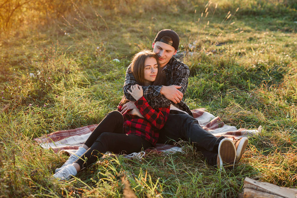 Young man hugs tenderly his pretty girlfriend. Young couple sits on the plaid at green lawn at sunset. - Valokuva, kuva