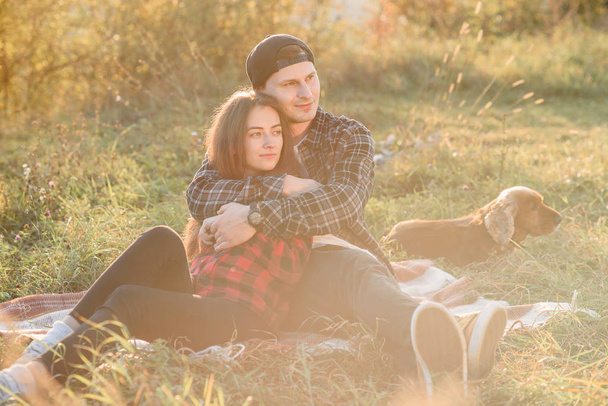 Young man hugs tenderly his pretty girlfriend. Young couple sits on the plaid at green lawn at sunset. - Foto, imagen