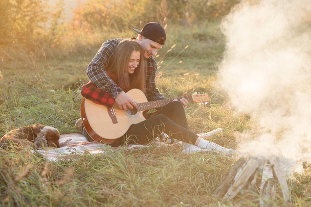 Young man playing on the guitar for his lovely girlfriend. Happy couple with guitar resting on picnic in spring park. - Foto, afbeelding