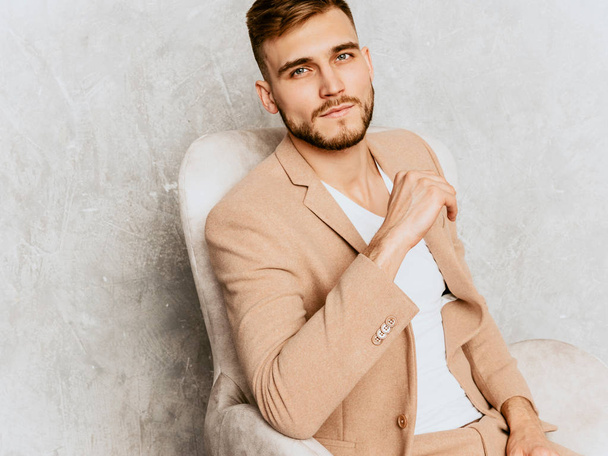 Portrait of handsome smiling hipster lumbersexual businessman model wearing casual beige suit. Fashion stylish man posing against gray wall sitting in chair in interior - Foto, immagini