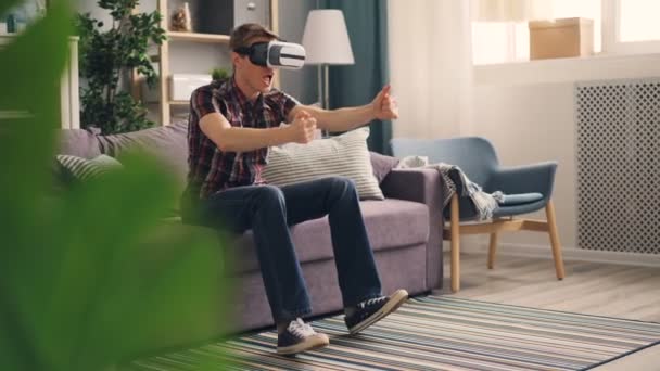 Excited guy is having fun with augmented reality glasses wearing headset and playing racing game moving hands and legs expressing positive emotions. - Felvétel, videó