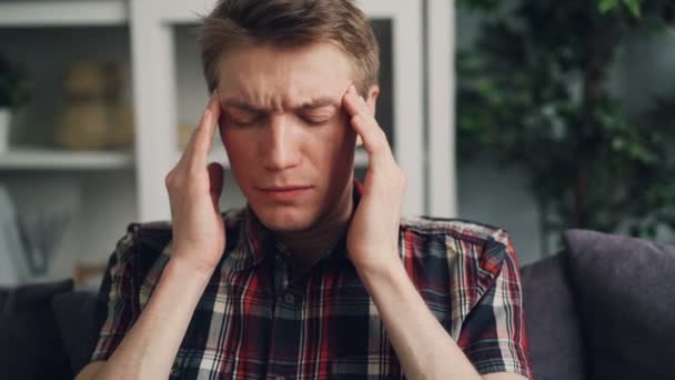 Unhappy sick young man is suffering from severe headache touching his head massaging temples indoors at home. Pain, tired youth and bad health concept. - Filmagem, Vídeo