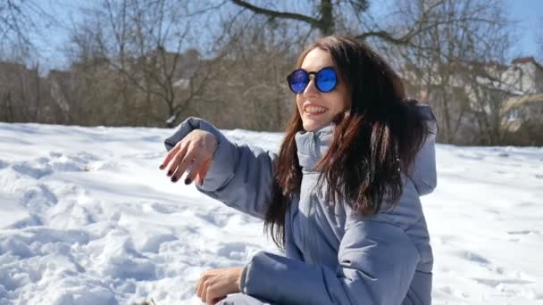 Hermosa morena en gafas de sol de espejo azul y pelo liso lanzando bolas de nieve jugando con alguien al aire libre en el parque por la mañana durante el día soleado pero frío a principios de primavera
 - Imágenes, Vídeo