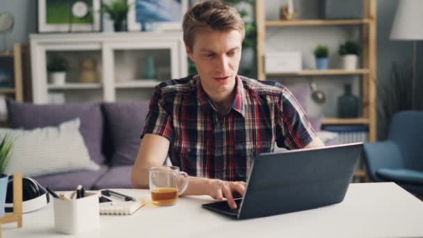 Successful male freelancer is working at home using laptop sitting at desk in studio looking at screen typing and smiling. Business and youth concept. - Metraje, vídeo
