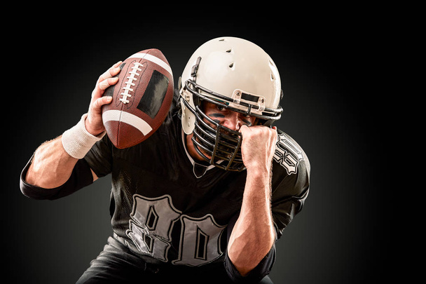 Giocatore di football americano in uniforme scura con la palla si prepara ad attaccare su uno sfondo nero
. - Foto, immagini