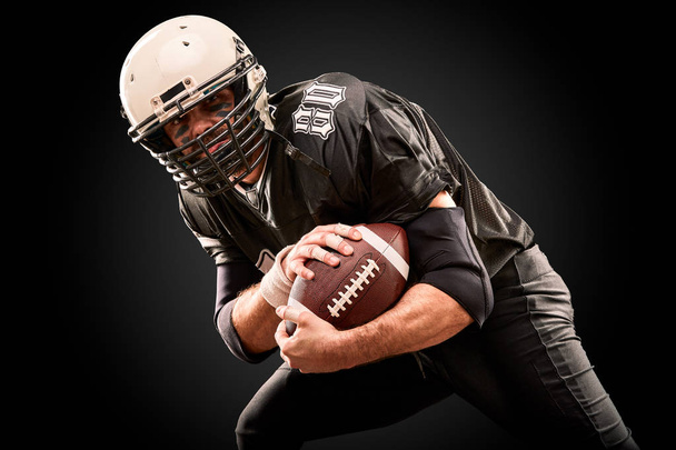 Giocatore di football americano in uniforme scura con la palla si prepara ad attaccare su uno sfondo nero
. - Foto, immagini