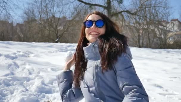Όμορφο κορίτσι σε μπλε γυαλιά ηλίου καθρέφτες και ίσια σκούρα μαλλιά, ρίχνοντας χιονόμπαλες παίζοντας με κάποιον σε εξωτερικούς χώρους στο πάρκο το πρωί κατά τη διάρκεια της ημέρας sunny αλλά κρύο νωρίς την άνοιξη - Πλάνα, βίντεο