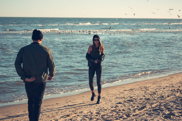 Young couple in love walks by the sea. Spring, autumn. The guy is wearing a jacket and hat. Girl in a hat and leather jacket with a scarf - Foto, Bild