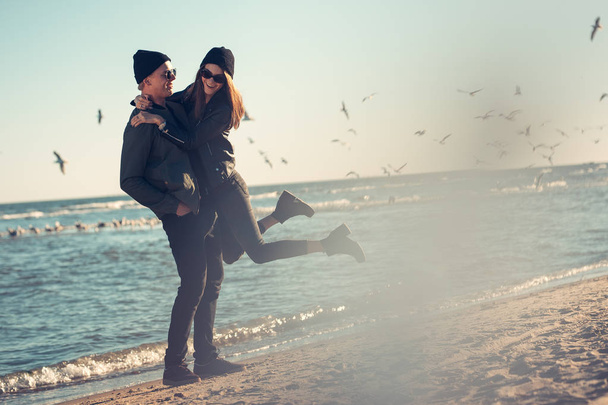 Νεαρό ζευγάρι με αγάπη βόλτες δίπλα στη θάλασσα. Άνοιξη, φθινόπωρο. Ο τύπος φοράει ένα σακάκι και καπέλο. Κορίτσι με ένα καπέλο και δερμάτινο σακάκι με ένα μαντήλι - Φωτογραφία, εικόνα