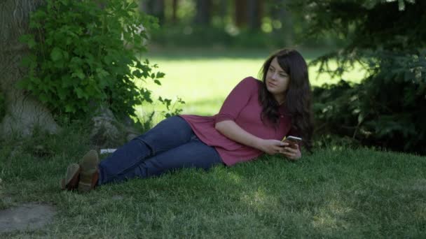 Young woman lying in the grass looing at her phone in a park enjoying the shade. - Imágenes, Vídeo